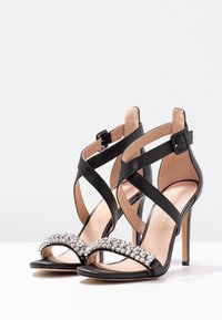 Mustat korkokengät, joissa on ristikkäiset nilkkahihnat ja strassisomisteet varvasremmissä, valkoista taustaa vasten.