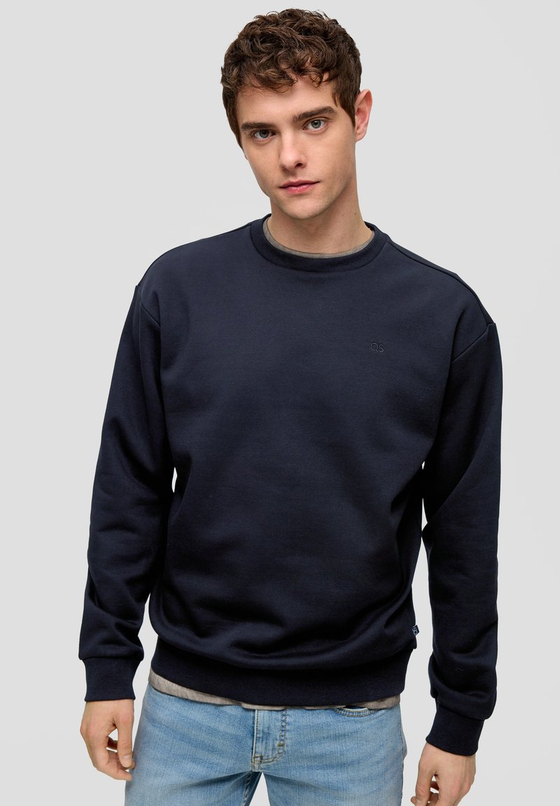 Felpa navy in misto cotone, con collo a girocollo e polsini a coste, dotata di un piccolo logo sul petto. Indossata sopra una maglietta grigia.