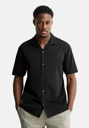 Schwarzes Kurzarmhemd mit einem breiten Kragen, vorne mit Knöpfen versehen und glatter Textur, kombiniert mit hellkarierten Hosen.
