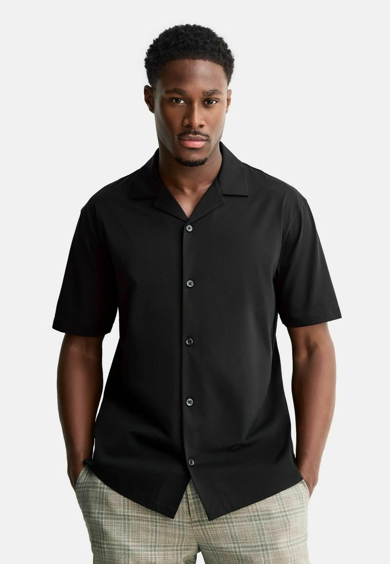 Črna kratka majica s širokim ovratnikom, z gumbi spredaj in gladko teksturo, kombinirana s svetlim karo hlačami.