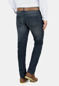 Muž oblečený v tmavě modrých džínách, hnědém pásku, bílém tričku a hnědých semišových botách, stojící s jednou nohou mírně zdviženou.