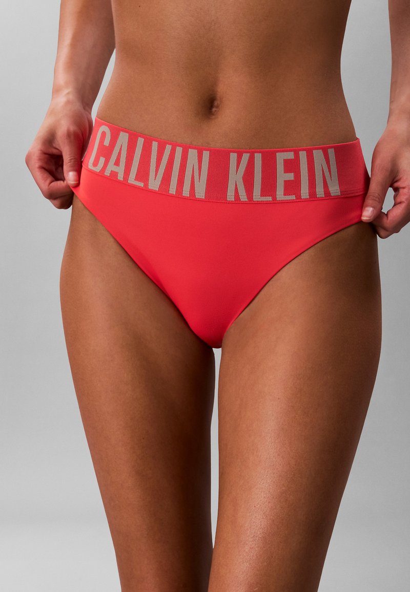 Kobieta ubrana w koralowe bikini Calvin Klein z szerokim paskiem z logo, trzymająca boki majtek.