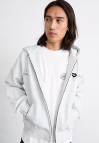 Felpa con zip grigia chiara, dotata di cappuccio e lacci, tasche frontali, polsini e orlo a coste. Presenta un logo a forma di cuore nero sul petto. Indossata sopra una t-shirt bianca.
