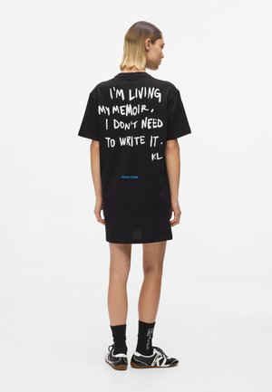 Abito t-shirt nero oversize con testo bianco sul retro con uno slogan. Include un dettaglio a hashtag blu sull'orlo.