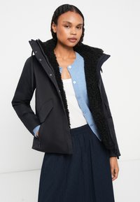 Giacca imbottita nera con foderatura in pelliccia sintetica nera, dotata di cappuccio, tasche laterali e zip. Indossata sopra un cardigan blu e una maglietta bianca.