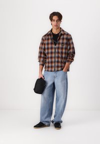 Brown and orange plaid zip-up shirt, black t-shirt, loose light blue jeans, black shoes, holding a small black bag.