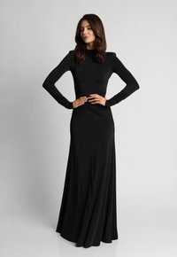 Kobieta w długiej czarnej sukni z marszczeniem w talii i rękawami stoi z rękami na biodrach, lekko spoglądając na bok na jednolitym tle.