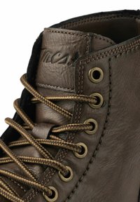 Bota de cuero marrón con ojales de bronce, con cordones trenzados marrones y costuras visibles. Superficie texturizada y logotipo grabado en el collar.