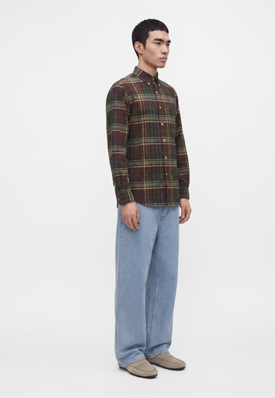 Homme debout portant une chemise à carreaux marron et verte boutonnée, un jean bleu clair à jambes larges et des chaussures beige à enfiler sur un fond blanc.