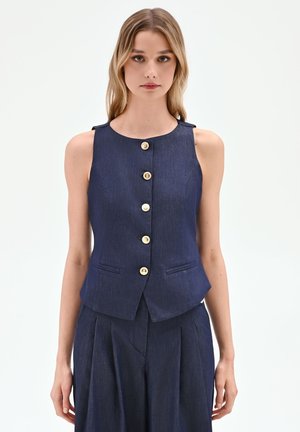 Donna che indossa un gilet smanicato blu navy con bottoni e pantaloni plissettati abbinati su sfondo uniforme.