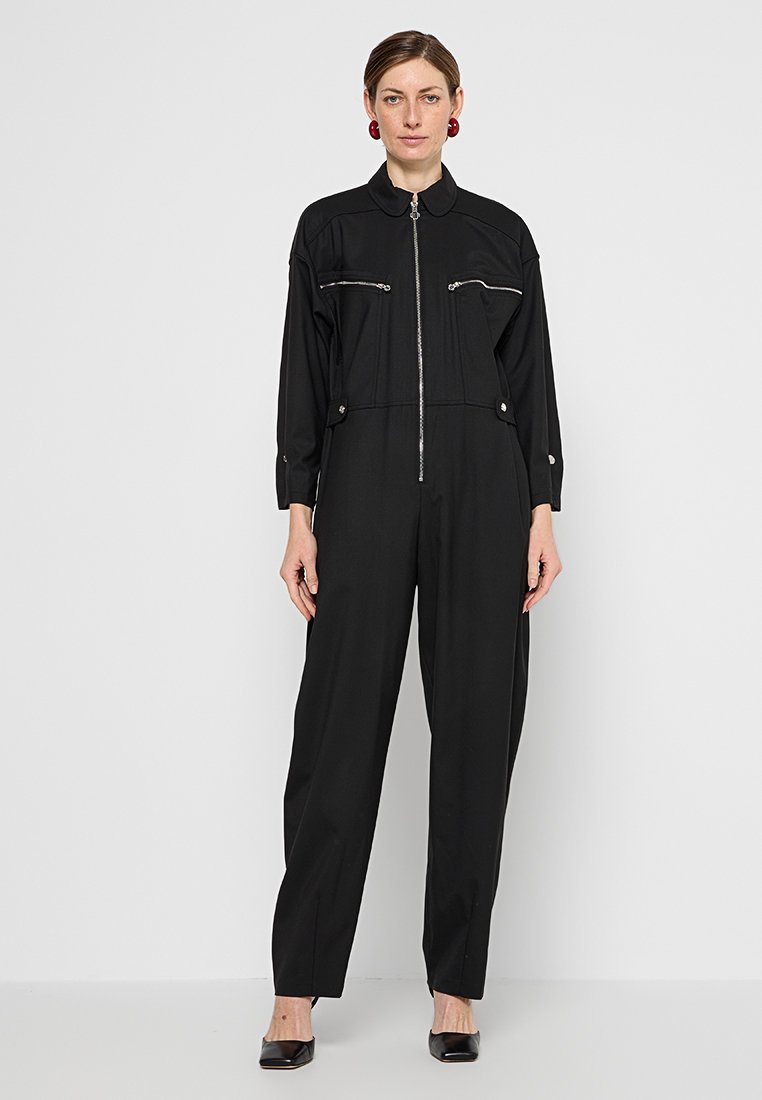 Schwarzer Overall mit Reißverschluss vorne, umklappbarem Kragen und zwei Brusttaschen. Er hat lange Ärmel und schmal zulaufende Beine. Glatte Stofftextur.