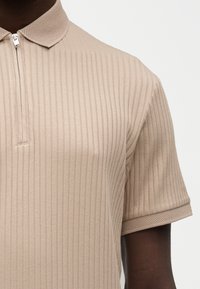 Gros plan sur une personne à la peau foncée portant un polo beige à manches courtes côtelé avec une fermeture éclair, un col texturé et des bords de manches texturés.