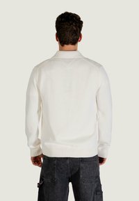 Biały dziany sweter z kołnierzem, z prążkowanymi mankietami i krawędzią. Widok od tyłu pokazuje prosty fason i subtelną fakturę. Noszony z ciemnymi spodniami.