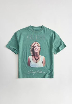 Zielony t-shirt z krótkim rękawem z uśmiechniętą kobietą w białym topie i naszyjniku, z tekstem „GUESS” u góry i podpisem odręcznym kursywą poniżej.