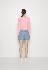 Różowa bluzka z długim rękawem w geometryczny wzór, zestawiona z niebieskimi szortami dżinsowymi. Model nosi białe trampki i stoi na jednolitym tle.