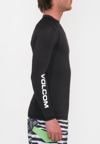 Czarna koszulka rash guard z długim rękawem i białym logo "VOLCOM" na rękawie, w połączeniu z kąpielówkami w paski. Model trzyma zielony przedmiot.
