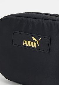 Czarny nylonowy worek z złotą naszywką logo Pumy, o gładkiej fakturze i zamknięciu na zamek. Okrągły kształt z minimalnymi elementami designu.