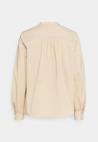 Beige bomullblus, långa ärmar med knäppte manschetter, detaljer på ryggen och lätt rynkad tyg för extra textur.