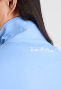 Light blue, soft fabric polo with a high collar. Features white embroidered text "Vivere Per Sempre" on the back near the neckline.