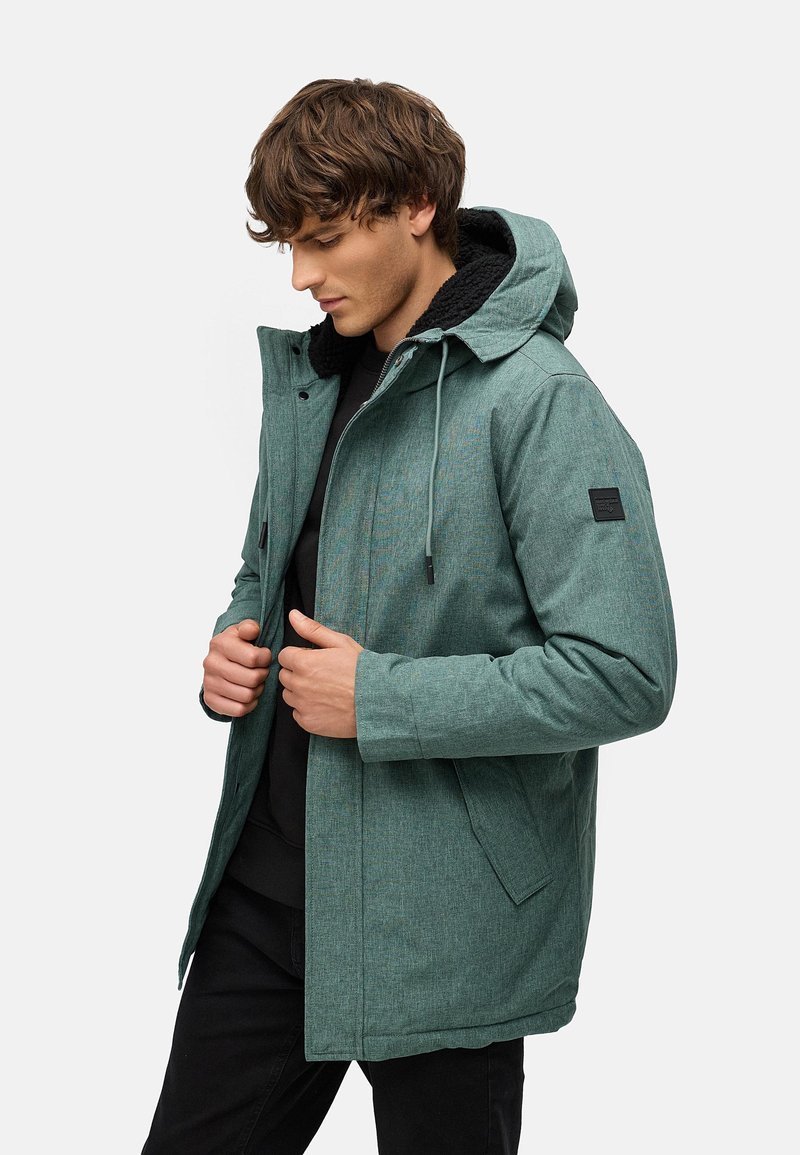 Chaqueta verde texturizada con capucha, con un forro de sherpa negro y un parche lateral. El modelo abrocha la chaqueta, de pie frente a un fondo uniforme.