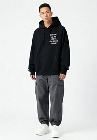 Felpa nera con cappuccio e testo bianco, tasca frontale a marsupio, abbinata a pantaloni cargo grigi e sneakers bianche. Vestibilità e design casual.