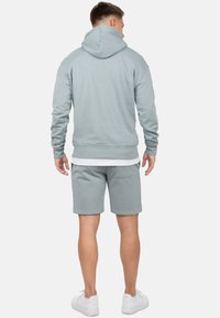 Lichtblauwe sweatshirt met een capuchon en lange mouwen, gecombineerd met bijpassende korte broek. Het materiaal lijkt zacht en textuur te hebben. Witte sneakers worden gedragen.