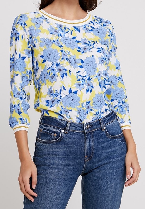 Vrouw in een geel-witte bloemenblouse met blauwe bloemen en driekwart mouwen, gecombineerd met blauwe denim jeans.