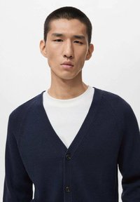 Navy ribbed cardigan with a V-neck, featuring a button closure and contrasting white T-shirt underneath. Soft texture, lightweight fabric.
