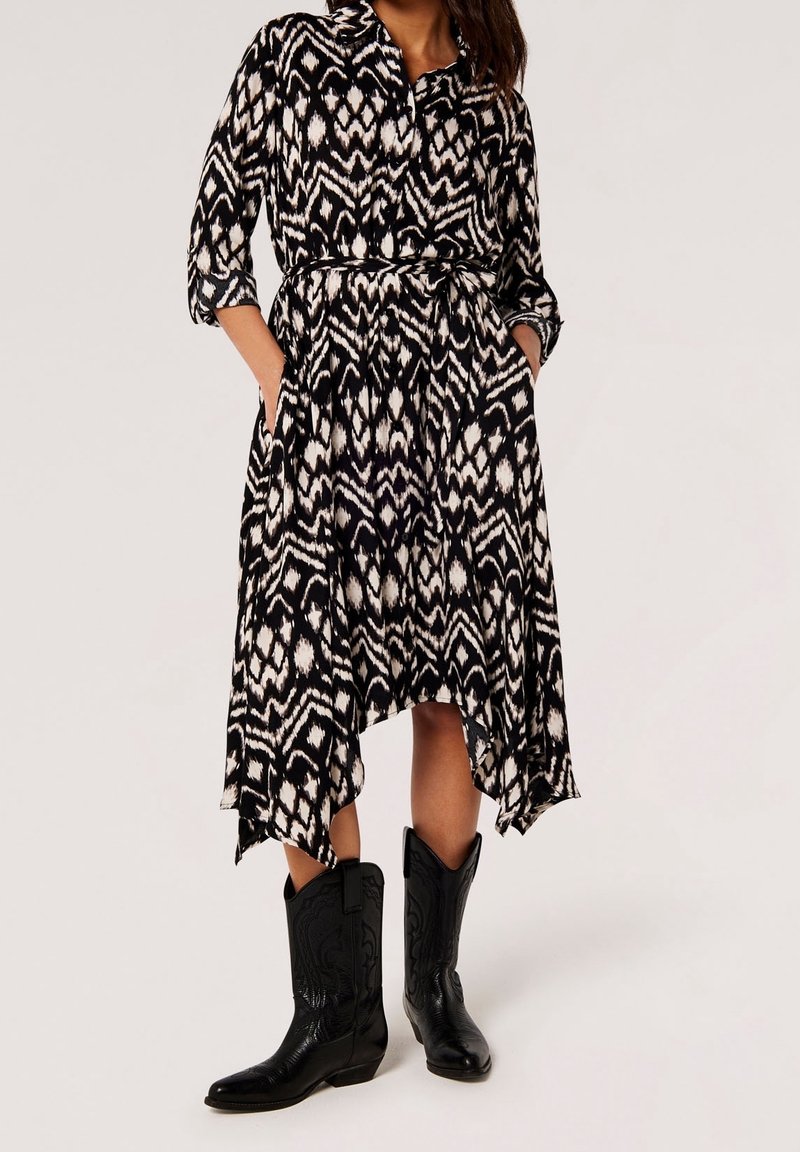 Femme portant une robe midi à motif noir et blanc avec des manches trois-quarts et des bottes de cow-boy noires, mains dans les poches de la robe.