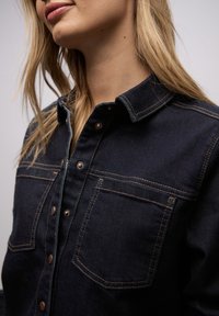 Gros plan sur une personne portant une veste en denim foncé avec des coutures marron et des boutons-pression en métal, montrant le col et les poches poitrine.