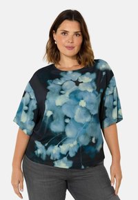 Blommig blus i nyanser av blått och svart, korta ärmar, rund halsringning, mjukt tyg med en len textur, bärs med grå byxor.