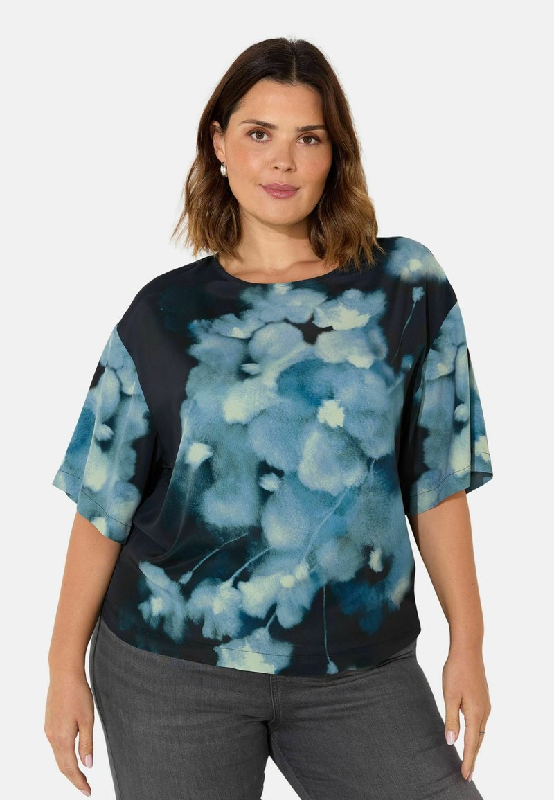 Blommig blus i nyanser av blått och svart, korta ärmar, rund halsringning, mjukt tyg med en len textur, bärs med grå byxor.