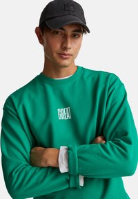 Jeune homme portant une casquette noire et un sweat-shirt vert avec le texte "GREAT", les bras croisés, et une manche de chemise blanche qui dépasse.
