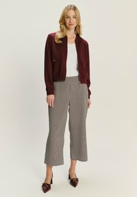 Bordeaux bomberjack met voorkeurszakken, witte top en gemusterde cropped broek in een beige en bruine geometrisch ontwerp; bruine puntige hakken.