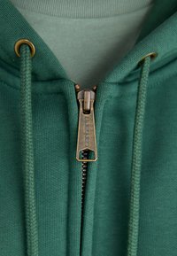 Grüne Kapuzenjacke mit einem metallischen Reißverschluss, strukturiertem Stoff mit gerippten Kanten und Zugbändern mit metallischen Ösen. Die innere Schicht ist in einem helleren Grün gehalten.