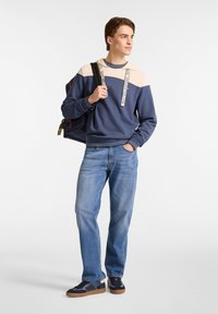 Sudadera azul marino con detalles en crema y correas con logo, combinada con jeans azul claro y zapatillas marinas. Mochila colgada de un hombro.