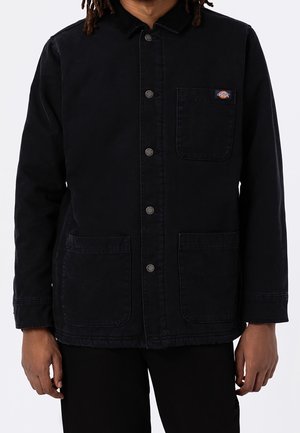 Mann trägt eine schwarze Jacke mit Knöpfen, drei vorderen Taschen und einem kleinen bunten Logo-Patch auf der oberen linken Tasche.