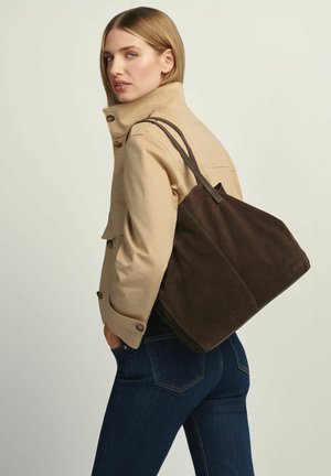 Kobieta w beżowej kurtce i ciemnych dżinsach niosąca dużą ciemnobrązową zamszową torbę na ramię, spoglądająca przez ramię wstecz.