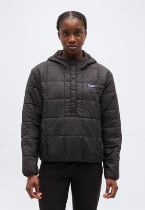 Žena v čiernej prešívanej bundi s kapucňou a logom Patagonia, stojaca proti jednolitému svetlému pozadiu, otočená čelom.