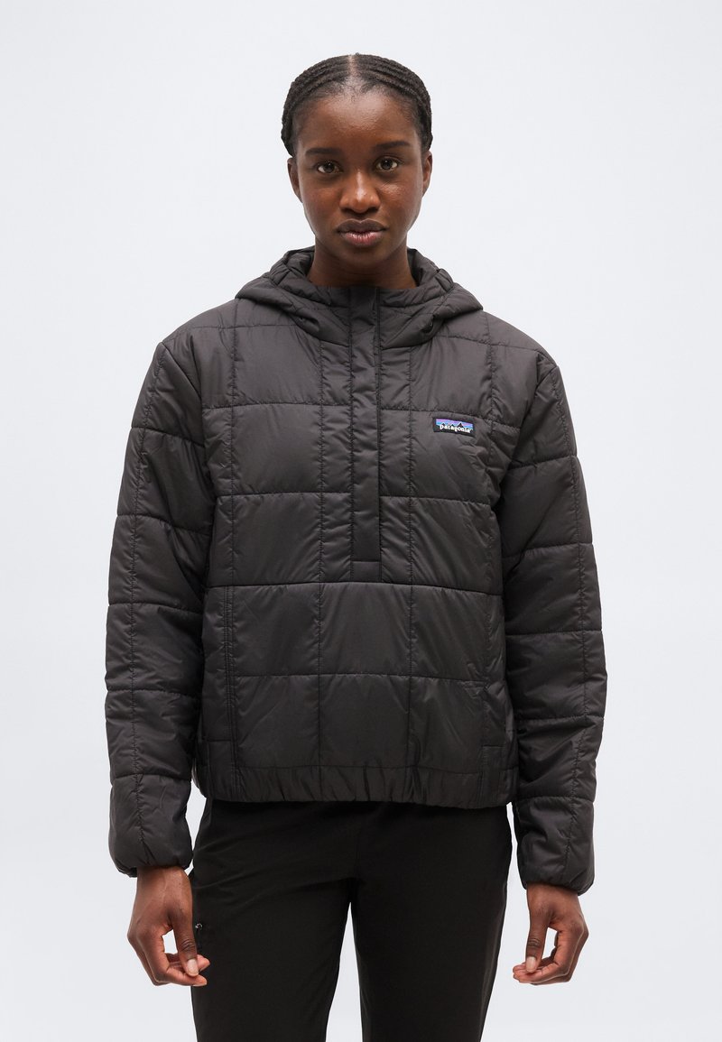 Donna con giacca nera trapuntata con cappuccio e logo Patagonia, in piedi davanti a uno sfondo chiaro uniforme, rivolta in avanti.