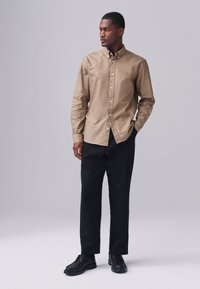 Overhemd met knopen in beige stof, met lange mouwen en een normale pasvorm. Gecombineerd met zwarte broek en zwarte veterschoenen.