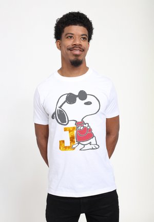 Mand iført hvid T-shirt med sort og rød Snoopy grafik og glitrende gyldent bogstav J, står med hænderne bag ryggen og smiler let.