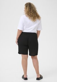 Shorts noirs à taille élastique avec une coupe décontractée, associés à un t-shirt blanc à manches courtes. Le mannequin a des cheveux doux et bouclés.