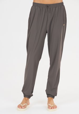 Persoon draagt losse, donkergrijze joggingbroek met elastische taille en enkels, gecombineerd met lichtgekleurde slippers, staand op een witte achtergrond.