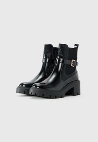 Bottines en cuir noir avec talons épais, panneaux élastiques sur les côtés, motifs embossés et brides à boucle dorée, présentées sur un fond blanc.