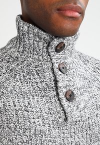 Pull gris en tricot avec un col montant, présentant trois boutons marron et un motif texturé. Gros plan mettant en valeur les détails du tissu.