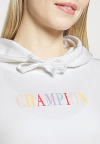Biely mikina s kapucňou na sťahovanie, so multicolor nápisom "CHAMPION" naprieč hrudníkom s prechodom farieb. Mäkká textúra látky.