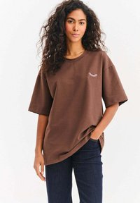 Žena s kudrnatými vlasy, oblečená v volném hnědém tričku s malým bílým motivem řas, tmavě modré džíny, stojí s jednou rukou v kapse.