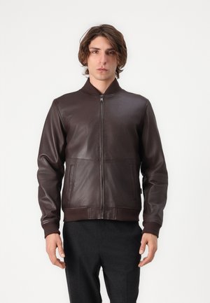 Junger Mann trägt eine dunkelbraune Lederjacke mit Reißverschluss, gerippten Bündchen und Kragen, steht vor einem schlichten weißen Hintergrund.