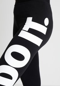 Czarne legginsy z dużym białym, wyraźnym napisem "doll.i" nadrukowanym diagonalnie na udzie i biodrze.